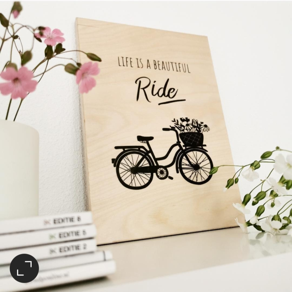 Afbeelding van een fiets met de tekst "life is a beautiful ride" geprint op een houten plankje naast bloemen en een stapel magazines.