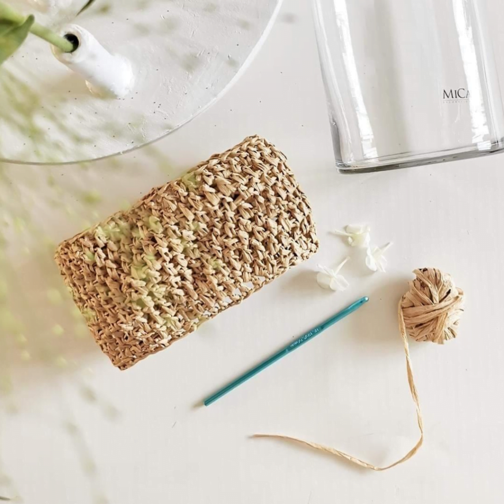 Haken met raffia in progress, met een haaknaald en bolletje raffia op tafel voor een vaas.