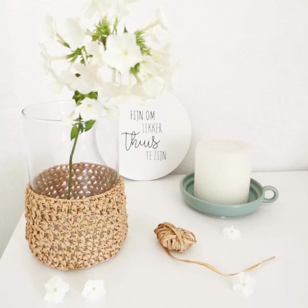 Doorzichtige vaas met een bloem, met daar omheen raffia gehaakt op een tafeltje, met een kaars, een sier bordje en een bolletje raffia wat over is van het haken.