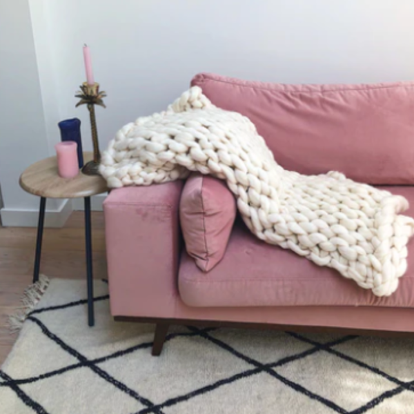 Een ivoorkleurige gebreide deken van lontwol op een roze bank, met een tafeltje met roze kaarsen en een woonkleed.