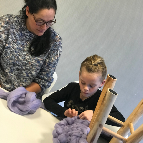 Liz van 7 leert van docent Ratna van September18.nl hoe ze de hoes voor een krukje moet breien met haar handen met lila lontwol.