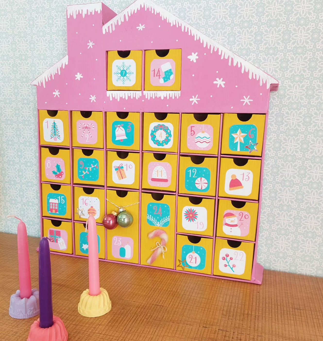 Advent huis geknutseld door Vrolijk Haakmeisje met felle kleuren geel, groen en roze, met kaarsen ervoor op tafel.