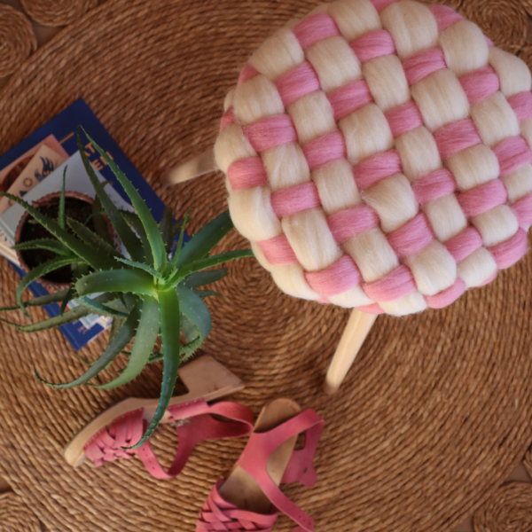 Boven aanzicht van een geweven patroon met roze en ivoor wol om een melkkruk heen, met schonen en een plant als styling ernaast.