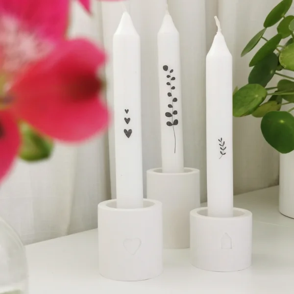 3 witte kaarsen versierd met zwarte stickers in zelf gegoten witte kaarsenhouders op een witte tafel met bloemen.