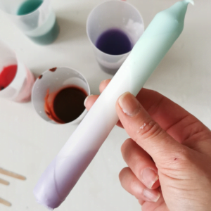 Voorbeeld van een gedipte verf kaars in mint groen en lila met op de achtergrond bekers met geverfde soya wax.